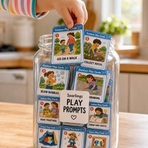 May include: A clear glass jar filled with illustrated play prompt cards. The cards feature colorful images and text like "Go on a Walk" and "Draw and Create." A child's hand reaches for a card. The jar also has a white label that says "Soarlings Play Prompts."
