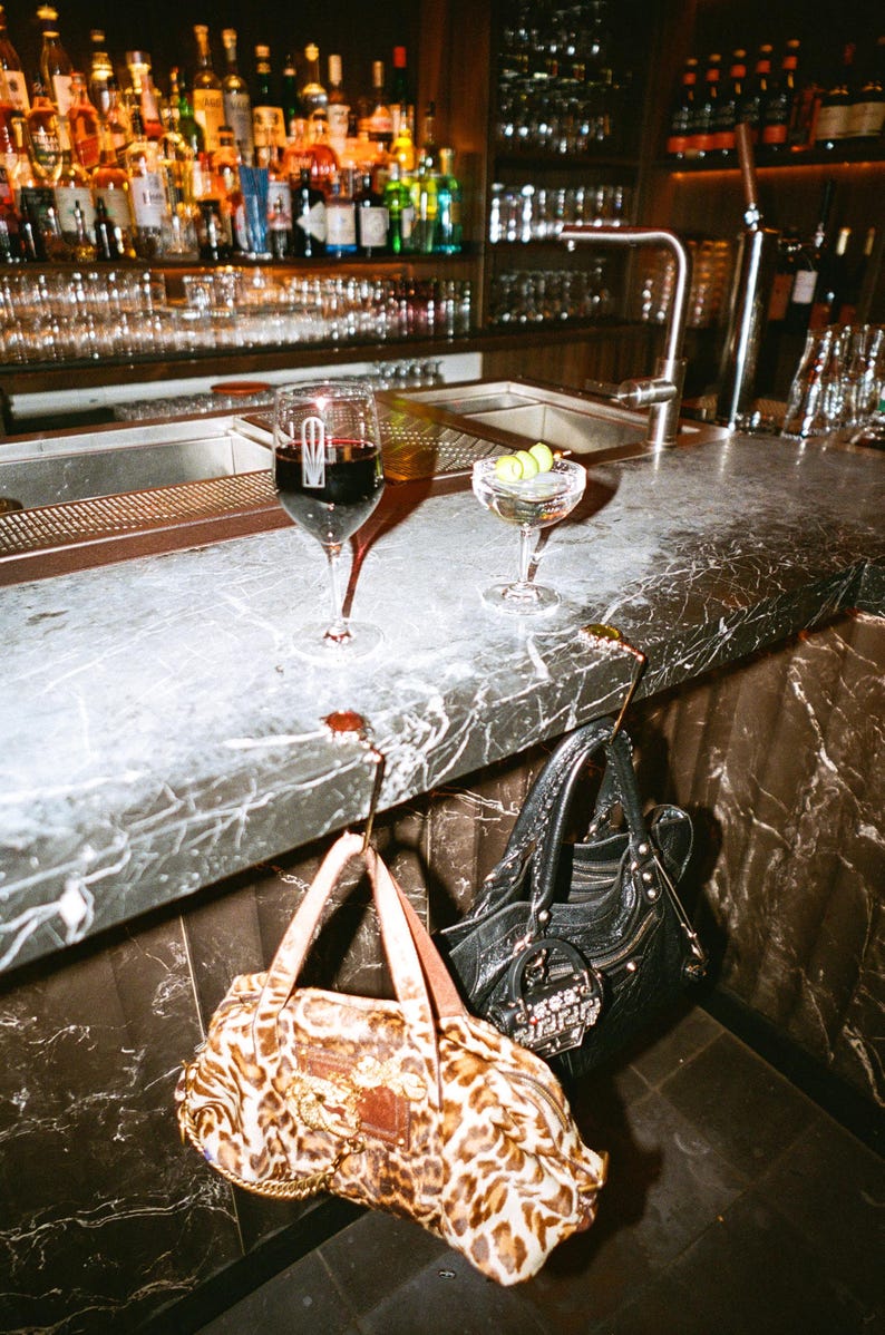 Puede incluir: Un bolso con estampado de leopardo con asas de cuero marr&oacute;n y un bolso de cuero negro con herrajes plateados, ambos colgados de ganchos met&aacute;licos en el borde de una barra de m&aacute;rmol gris con bebidas colocadas encima.