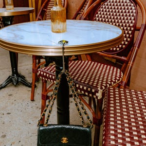 Puede incluir: Un bolso de mano de cuero acolchado negro con correa de cadena dorada colgado de un gancho de metal plateado sujeto a una mesa redonda con tapa de m&aacute;rmol blanco. El bolso presenta un cierre con logotipo dorado en la solapa y cadena entrelazada.