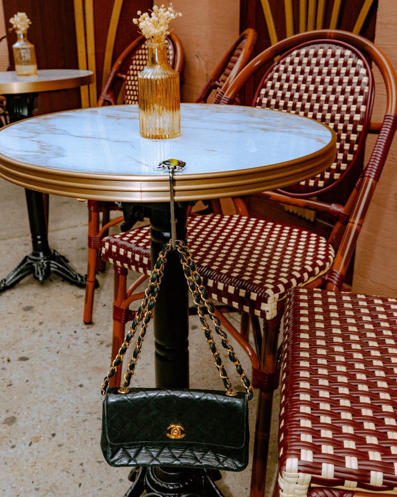 Puede incluir: Un bolso de mano de cuero acolchado negro con correa de cadena dorada colgado de un gancho de metal plateado sujeto a una mesa redonda con tapa de m&aacute;rmol blanco. El bolso presenta un cierre con logotipo dorado en la solapa y cadena entrelazada.