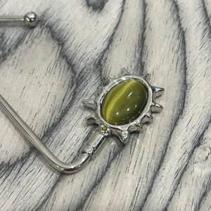May include: A silver-coloured handbag hook with a decorative sunburst design. The sunburst has an oval, olive-green stone at its centre, with a small green accent stone. The hook is designed to hang from a table or counter.