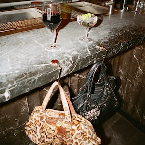 May include: Two metal purse hooks are attached to the edge of a marble bar counter. A leopard print handbag with gold hardware hangs from one hook, while a black leather handbag with silver studs and a small charm hangs from the other hook.