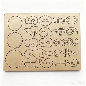 May include: A rectangular light-colored wooden stencil sheet featuring laser-cut designs. The board includes three circular shapes on the left and a grid of numbers from zero to nine arranged in three columns across the remaining surface area.