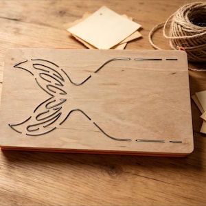 Puede incluir: Un troquel de madera con un diseño de cola de sirena. El troquel es de color marrón claro con un borde rojo y detalles metálicos. También hay un carrete de hilo y varias etiquetas pequeñas en blanco.
