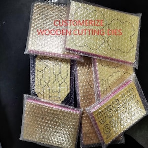Könnte beinhalten: Eine Sammlung von Holzstanzformen in verschiedenen Formen, darunter Schleifen, Blumen und Zahlen, verpackt in Luftpolsterfolie. Der Text "CUSTOMIZE WOODEN CUTTING DIES" ist auf der Verpackung zu sehen. Die Stanzformen sind wahrscheinlich für Bastel- oder Scrapbooking-Projekte.