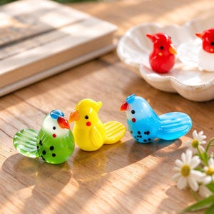May include: Five small glass bird figurines in various colours including green, yellow, blue, red, and white. Each bird features a rounded body, a small beak, and a flat tail. The green and blue birds have black spots on their bodies.