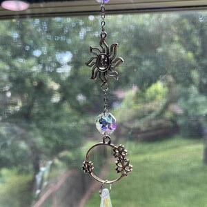 May include: A decorative suncatcher with a silver sun charm, a clear crystal bead, a silver ring with flower accents, and a teardrop-shaped crystal pendant. The suncatcher hangs from a chain and is suspended in front of a window.