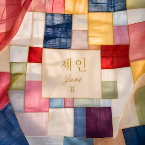 Könnte beinhalten: Ein Patchwork-Textil mit einer Vielzahl von Farben, darunter Rot, Blau, Gelb und Rosa. In der Mitte steht der Name "Jane" in goldener Schrift, darüber koreanische Schriftzeichen. Das Textil besteht aus zusammengenähten Quadraten.