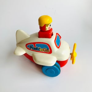 May include: A white and red toy airplane with a yellow propeller and a small figure of a pilot in the cockpit. The airplane has the words "Push n Go" printed on the side.