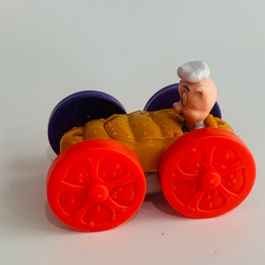May include: A toy car shaped like a pig with orange wheels and a yellow body. The pig is wearing a white sailor hat and has a pink face.
