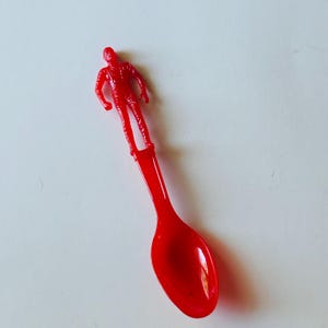May include: A red plastic spoon with a small red plastic figure of a person standing on the handle.