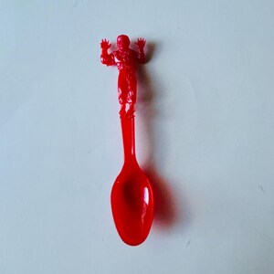 May include: A red plastic spoon with a red plastic figure of a person standing on the handle.