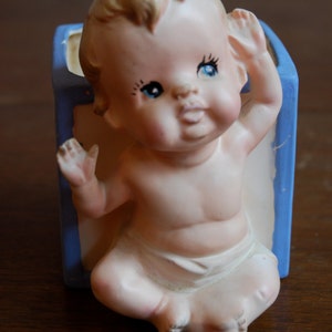 Puede incluir: Un macetero de cerámica vintage con forma de bebé que lleva un pañal. El macetero está pintado en tonos azules y blancos y presenta un bebé sonriente con ojos azules.
