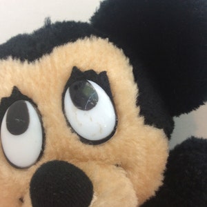 Puede incluir: Primer plano de un juguete de peluche de Mickey Mouse con ojos grandes y blancos y pupilas negras. El juguete tiene orejas negras y una cara marr&oacute;n.