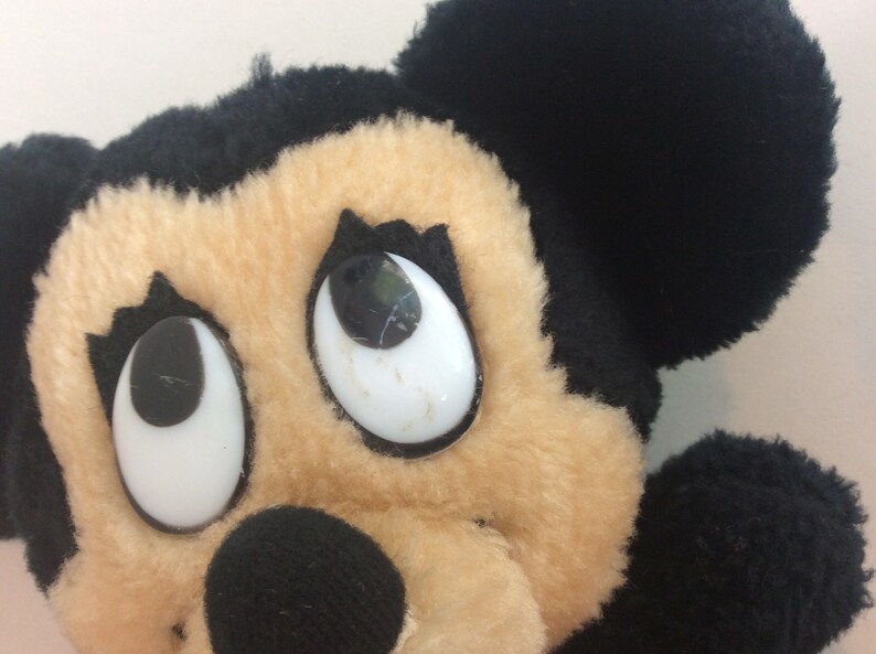 Puede incluir: Primer plano de un juguete de peluche de Mickey Mouse con ojos grandes y blancos y pupilas negras. El juguete tiene orejas negras y una cara marr&oacute;n.