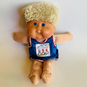 May include: A Cabbage Patch Kid doll wearing a blue and red basketball jersey with the text "Olympic Kids 1996 Official Team Mascot USA" and Olympic rings. The doll has blonde hair and blue eyes.