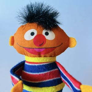 May include: A plush toy featuring a smiling orange face with a red nose, white eyes, and black hair. The toy wears a striped blue, red, and yellow sweater. The toy's design is reminiscent of a popular children's character.