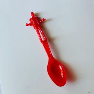 May include: A red plastic spoon with a small figure of a person standing on the handle.