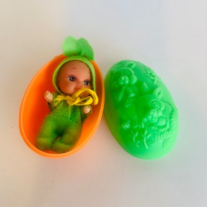 May include: A green and yellow baby doll in a green and white outfit is inside a bright orange plastic egg. The egg is open and the doll is looking out. The other half of the egg is green and has a design of two bunnies on it.