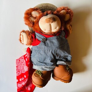 May include: A plush teddy bear wearing a straw hat, red shirt, and blue and white striped overalls. The bear has brown fur, a tan snout, and brown boots. A red paisley bandana is included.