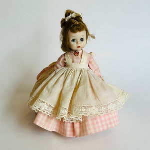 May include: A vintage porcelain doll wearing a white dress with a pink and white gingham underskirt and a lace trim. The doll has brown hair, blue eyes, and a red jewel necklace.