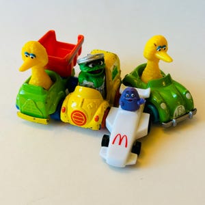 May include: A set of five Sesame Street character toy cars. The cars are yellow, green, and white with red, orange, and blue accents. The characters include Big Bird, Cookie Monster, Oscar the Grouch, and Elmo. The cars are in good condition and would be a great addition to any Sesame Street fan's collection.