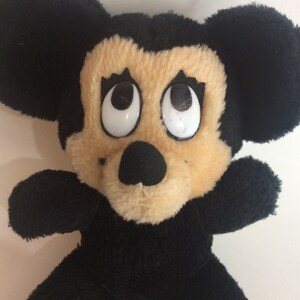Puede incluir: Un juguete de peluche Mickey Mouse negro y marr&oacute;n con grandes ojos blancos. El juguete tiene una nariz negra y orejas negras.