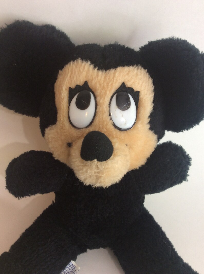 Puede incluir: Un juguete de peluche Mickey Mouse negro y marr&oacute;n con grandes ojos blancos. El juguete tiene una nariz negra y orejas negras.
