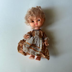 May include: A vintage doll with curly blonde hair and a brown dress with a floral pattern. The dress has a white apron with lace trim and orange accents. The doll has a sweet expression and is in good condition.