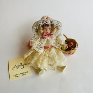 May include: A porcelain doll in a cream-colored lace dress and matching hat, adorned with pink accents. The doll holds a small woven basket filled with flowers. The tag reads "Jocelyn Mastrom".