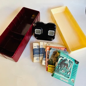 May include: A vintage View-Master viewer with a black plastic body and two red lenses. The viewer is surrounded by a collection of colorful View-Master reels featuring images of natural wonders, including a glacier and a sorrowful mother.