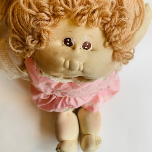 May include: A vintage Cabbage Patch Kid doll with blonde yarn hair, wearing a pink dress with white polka dots and lace trim. The doll has brown eyes and a painted smile.