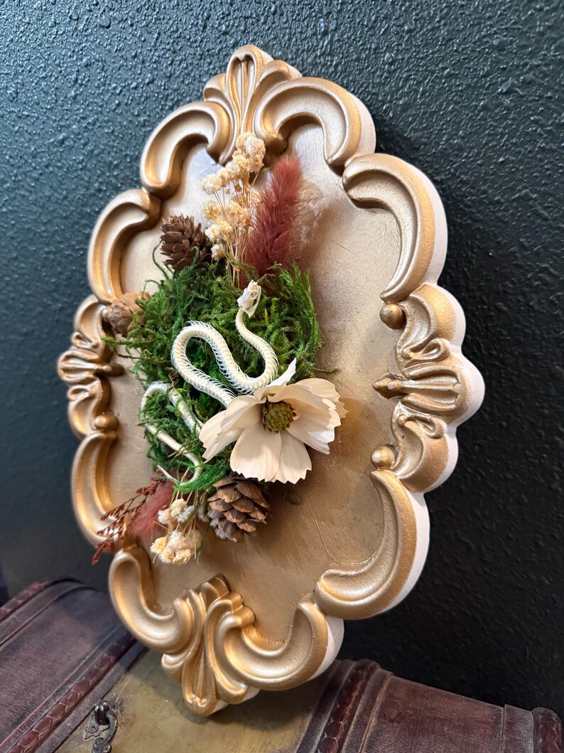 May include: A wall-mounted gold-painted ornate frame containing a white snake skeleton, green moss, a white flower, small pinecones, and dried botanical elements arranged in a cluster on a gold-colored backing surface.