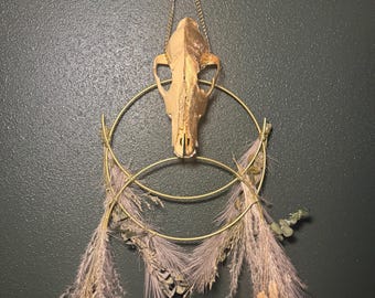 May include: A decorative wall hanging featuring a gold-painted animal skull suspended by a chain. The skull is centered within two gold hoops, adorned with dried, feathery, beige and lavender-colored grasses and foliage. The background is a dark green.