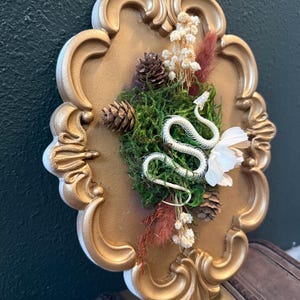 May include: A gold-painted, ornate oval frame containing a white snake skeleton, green moss, small pinecones, dried white flowers, and reddish-brown dried grasses. The arrangement is centered within the frame, creating a textured display.