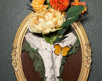 May include: An oval-shaped wall hanging with a gold frame. The artwork features a white bone structure, green moss, and a bouquet of orange and yellow flowers. A butterfly rests on the bone.