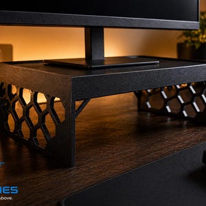 May include: Black monitor stand with a geometric honeycomb design on the sides. The stand is made of a dark material and is designed to elevate a computer monitor. The text "NJ SKIES On The Level Above" is visible.