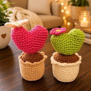 May include: Two crochet cactus plants. One has a pink heart-shaped cactus with a white flower, the other has a green heart-shaped cactus with a pink flower. Both are in tan and white pots with brown soil.