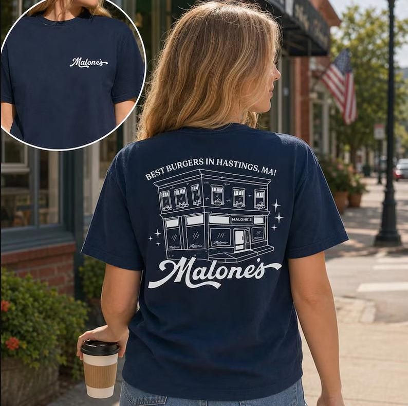 Op de afbeelding: Marineblauw t-shirt met de tekst "Malone's" op de voorkant en een gedetailleerde illustratie van een gebouw met de tekst "Best Burgers in Hastings, MA!" op de achterkant. Het shirt heeft een ronde hals.