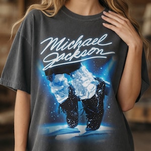 May include: Dark gray t-shirt featuring the name "Michael Jackson" in white script above an image of the singer's iconic dance pose. The graphic includes a blue and white starry background and the singer's black shoes.