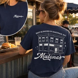 Op de afbeelding: Marineblauw t-shirt met het woord "Malone's" in witte letters op de voorkant. De achterkant toont een witte illustratie van een gebouw met de tekst "Best Burgers in Hastings, MA!" en de naam "Malone's".