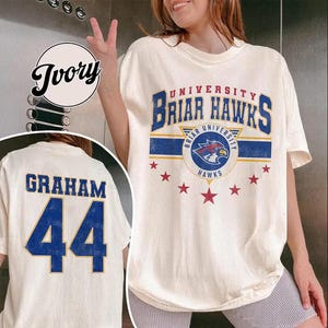 Può includere: T-shirt color crema con testo e logo "UNIVERSITY BRIAR HAWKS". Il davanti presenta un design blu e oro con stelle rosse. Il retro mostra "GRAHAM" e il numero "44" in blu.