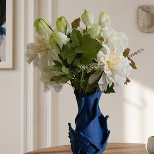Könnte beinhalten: Eine marineblaue Vase mit einem einzigartigen, geformten Design hält einen Strauß weißer Blumen und grünes Laub. Die Blumen umfassen große, offene Blüten und geschlossene Knospen. Die Vase steht auf einem Holztisch und erzeugt einen Kontrast aus Texturen und Farben.