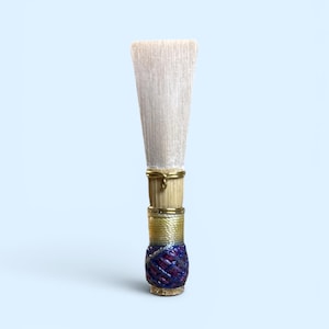 May include: A close-up of a reed for a woodwind instrument. The reed has a light-colored, rectangular tip and a decorative base with gold, blue, and purple accents. The background is a light blue.