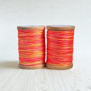May include: Two wooden spools wrapped with vibrant neon orange and yellow variegated thread. The thread features a high-contrast blend of bright fluorescent orange and sunny yellow tones wound tightly around the cylindrical wooden cores.