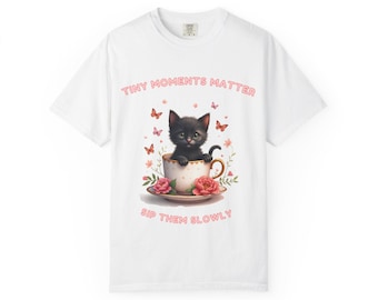 May include: White t-shirt featuring a black kitten in a teacup with pink roses and butterflies. The text "Tiny Moments Matter, Sip Them Slowly" is printed above and below the image. The shirt is a comfortable, casual style.