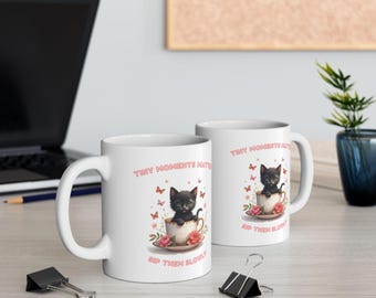 May include: Two white ceramic mugs with a whimsical design. Each mug features a black kitten sitting in a teacup, surrounded by butterflies and flowers. The mugs have the text "Tiny Moments Matter, Sip Them Slowly."