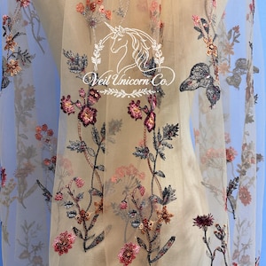 Pode incluir: Tecido bege transparente com bordados florais em tons de rosa, bordô e marrom. O design inclui flores delicadas, folhas e o texto "Veil Unicorn Co." em uma fonte decorativa. O tecido é provavelmente para véus ou roupas de noiva.