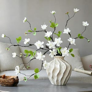 May include: A decorative off-white ceramic vase with a vertical ribbed texture holds a branch arrangement featuring delicate white flowers and small green leaves. The vase has a rounded body and a narrow neck, displaying a matte finish.
