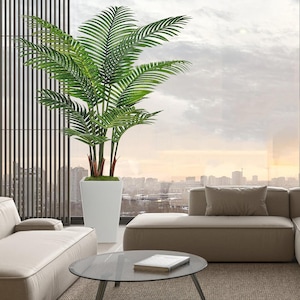 Puede incluir: Una gran palmera artificial con hojas verdes y tallos rojizos se exhibe en una maceta cuadrada blanca. La planta está en un salón moderno con un sofá de tonos neutros, una mesa de centro redonda de cristal y una vista de la ciudad.